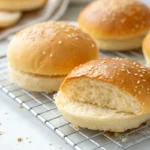 Image of freshly baked Egg White Burger Buns. The photo shows golden-brown, light-textured buns on a cooling rack, some sliced in half to show their airy insides. This highlights a healthier alternative for burger buns